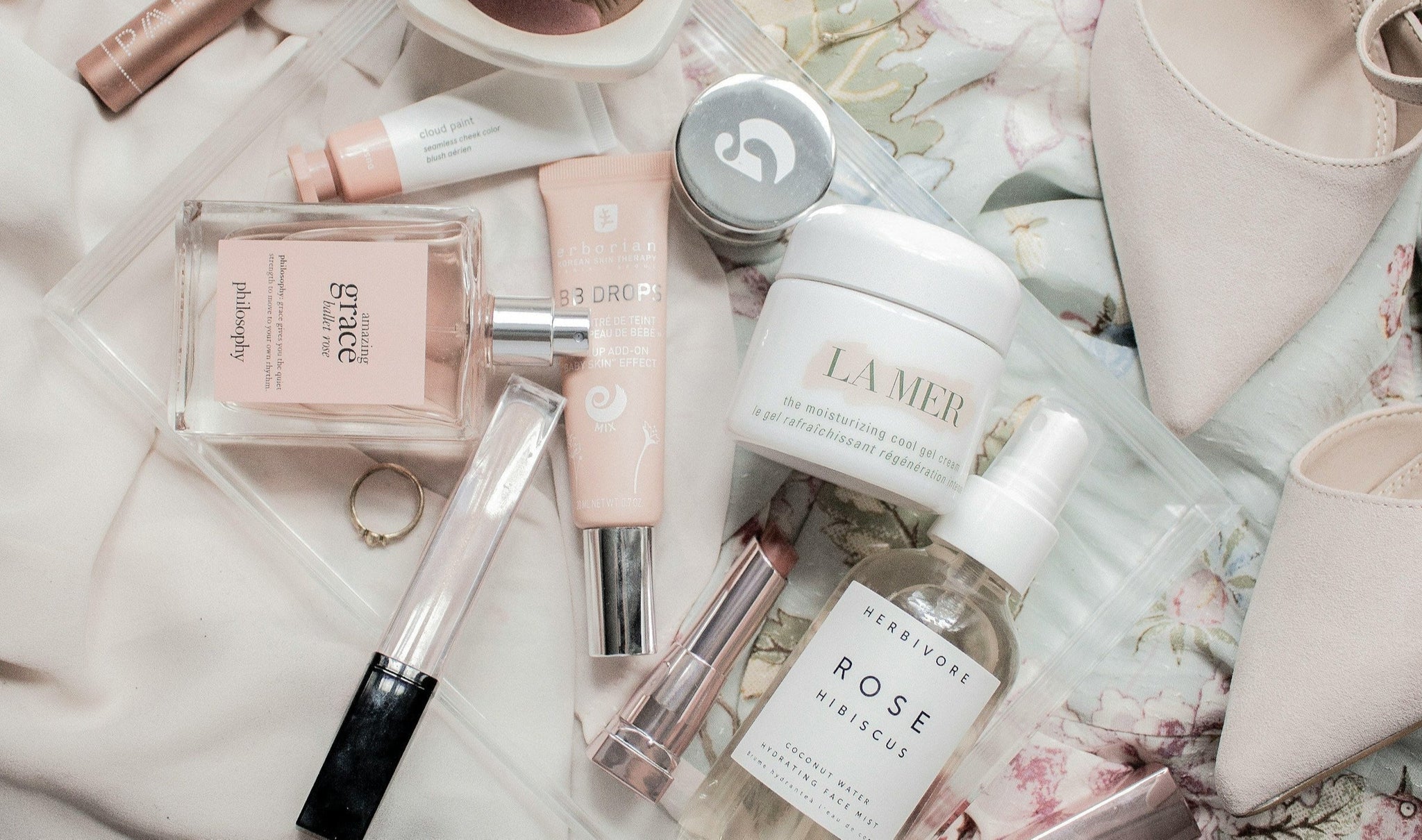 Collection of beauty products including a perfume bottle, lip gloss, and cream on a floral fabric background.